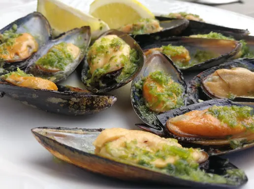 A plate of mussels in their shells topped with green mojo and herbs, with lemon wedges
