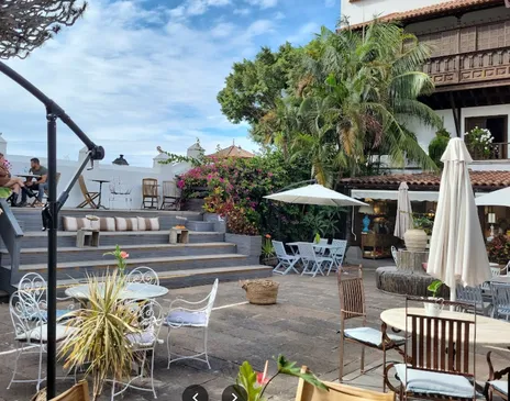 A leafy terrace with white wrought-iron tables, parasols and palms, beside a colonial building
