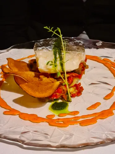A plated fillet of white fish resting on stewed peppers and vegetables, with sweet potato crisps and a swirl of orange sauce