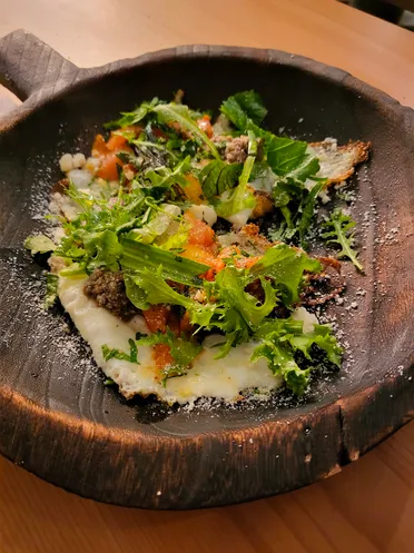 A plated dish on a dark wooden board: fish under dressed herb leaves, diced vegetables and dustings of powder