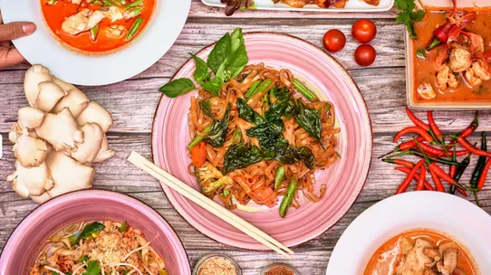 A Thai spread on a wooden table: pad thai on a pink plate, red curries, fresh chillies, oyster mushrooms and chopsticks
