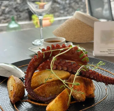 A plate of grilled octopus tentacle with roast potato wedges and herbs, on an outdoor table with a wine glass and a straw hat behind