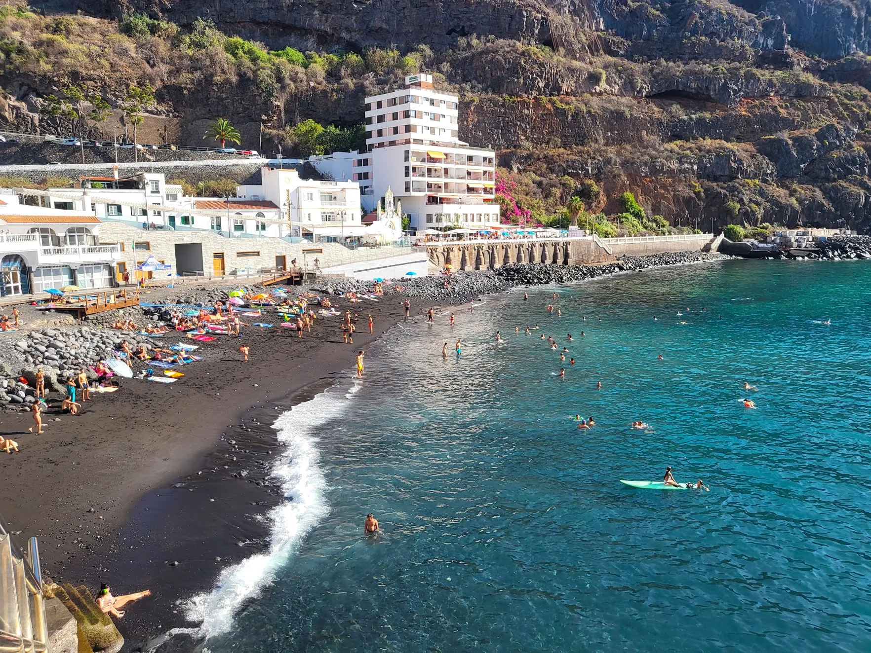 Playa de San Marcos from above: black sand, swimmers in turquoise water, and the white buildings and restaurant terraces along the seafront beneath the cliff