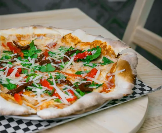 A thin-based pizza on a wooden board, topped with rocket, shaved cheese, roasted red peppers and cured meat